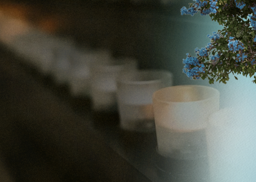 A softly lit, slightly grainy image showing a row of small translucent candle holders receding into the background on a dark surface, with the nearest candle in focus. On the right side, a cluster of small blue flowers and green leaves enters the frame, adding a gentle pop of color against the muted, dreamy atmosphere.